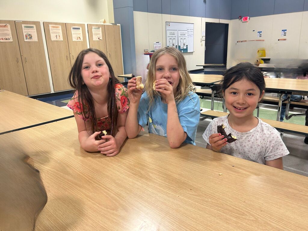 Three smiling girls, holding donuts in a cafeteria.