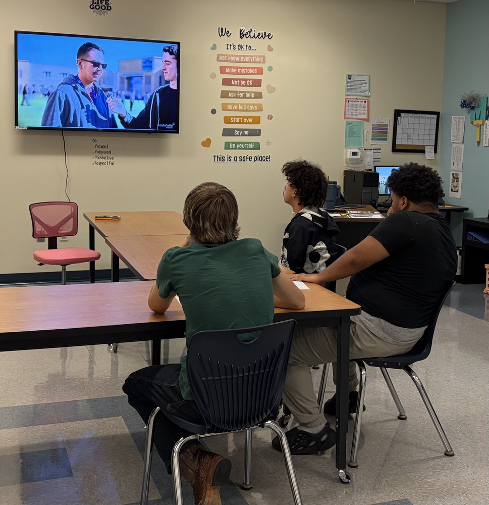 A group of 3 students sit in an classroom watching a television interview.