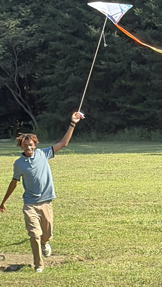Boy wearing a green shirt and khakis in a grassy field with trees in the background flies a homemade kite.