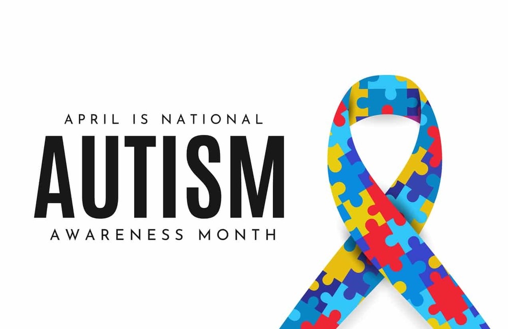 A graphic featuring a white background. On the right side is a large awareness ribbon filled with a multicolored puzzle piece pattern in blue, red, and yellow. To the left of the ribbon, the text reads: APRIL IS NATIONAL AUTISM AWARENESS MONTH. The word AUTISM is written in a large, bold, black sans-serif font.