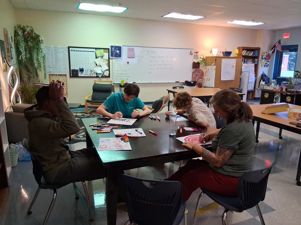 Classroom of 3 students and 1 teacher writing a mission statement at a large table.