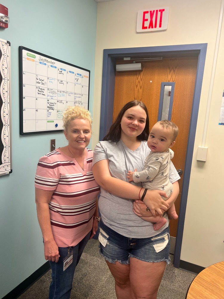 Two women pose for a picture. One is holding a baby. In an office like setting.