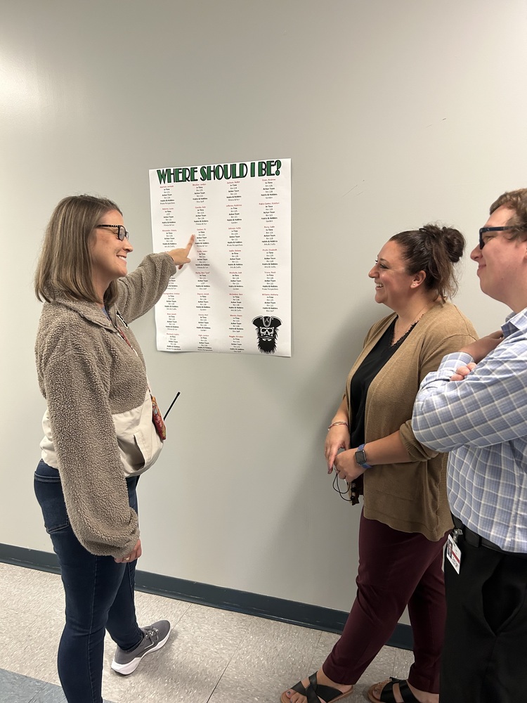 Two women and one man stand in a school hallway looking at a "Where Should I Be" poster on the wall.