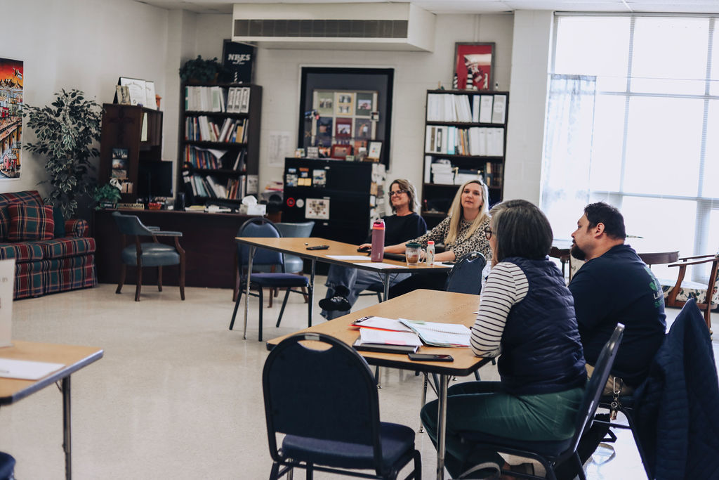 Educators sit around tables in a classroom setting, listening to a speaker off-camera during a professional learning meeting, with bookshelves and office furniture visible in the background.