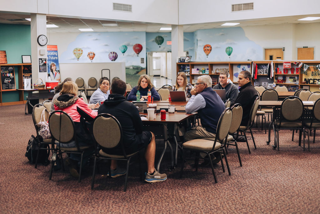 A small group of educators sit around a round table engaged in discussion during a meeting in a library or media center, with laptops, notebooks, and drinks on the table.