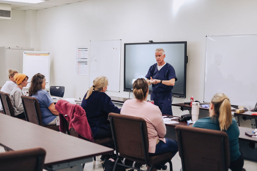 An instructor stands at the front of a classroom-style training room speaking to seated educators, who are facing forward and listening attentively during a professional learning session.