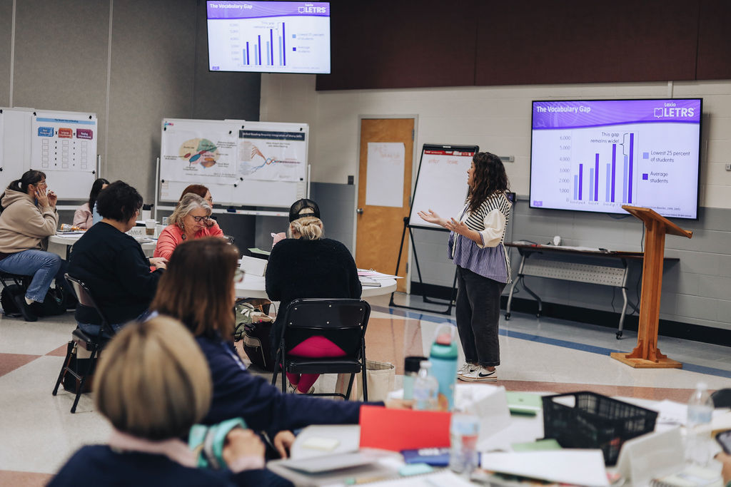 A facilitator leads a literacy-focused professional development session, gesturing toward a presentation screen showing data and charts while educators work and discuss at tables.