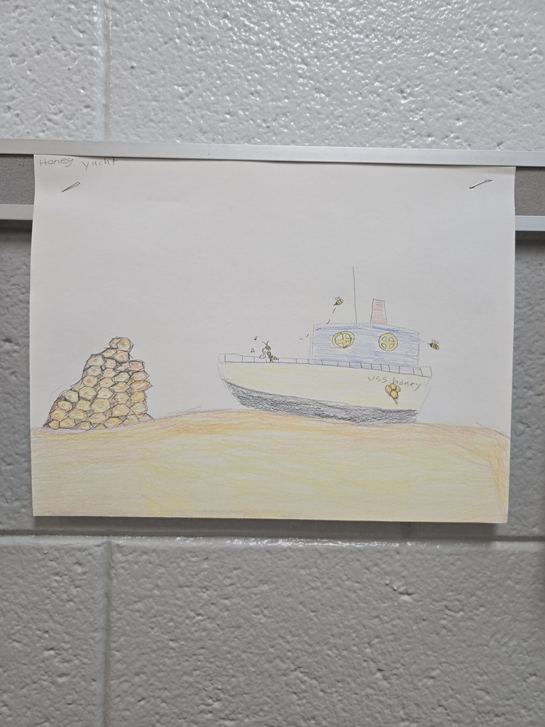 A student drawing taped to a classroom wall shows a boat labeled “USS Honey” floating on a golden honey-colored surface, with bees flying around it and a large honeycomb structure on the left. The illustration blends the words “honey” and “yacht” into a single creative scene.