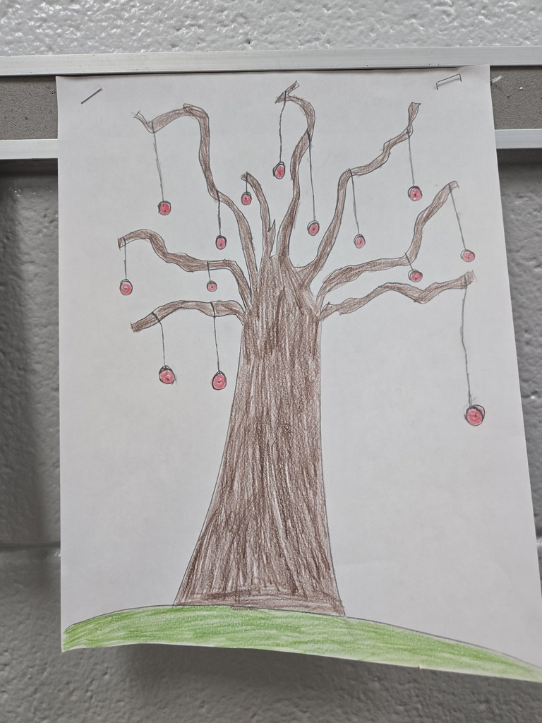 A student drawing taped to a classroom wall shows a large tree with multiple branches. Several yo-yos hang from the branches by strings, combining the ideas of a tree and yo-yo into one imaginative illustration.