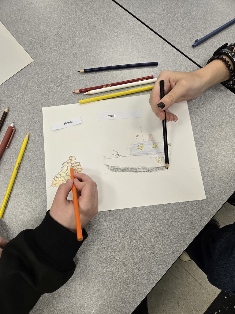 Two students sit at a classroom table coloring a shared drawing labeled “Honey” and “Yacht.” One student colors a honeycomb structure while the other shades a boat surrounded by bees. Colored pencils are spread across the table as the students work together on the artwork.