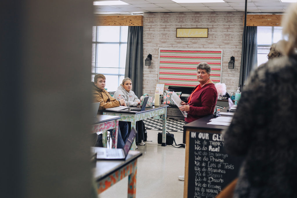 A substitute teacher stands at the front of a high school classroom holding a lesson plan while students sit at desks and look on, with classroom posters and learning materials visible in the background.