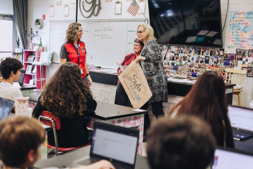 Two school staff members present a “January Sub of the Month” sign to a substitute teacher at the front of a classroom while students observe from their desks with laptops open.
