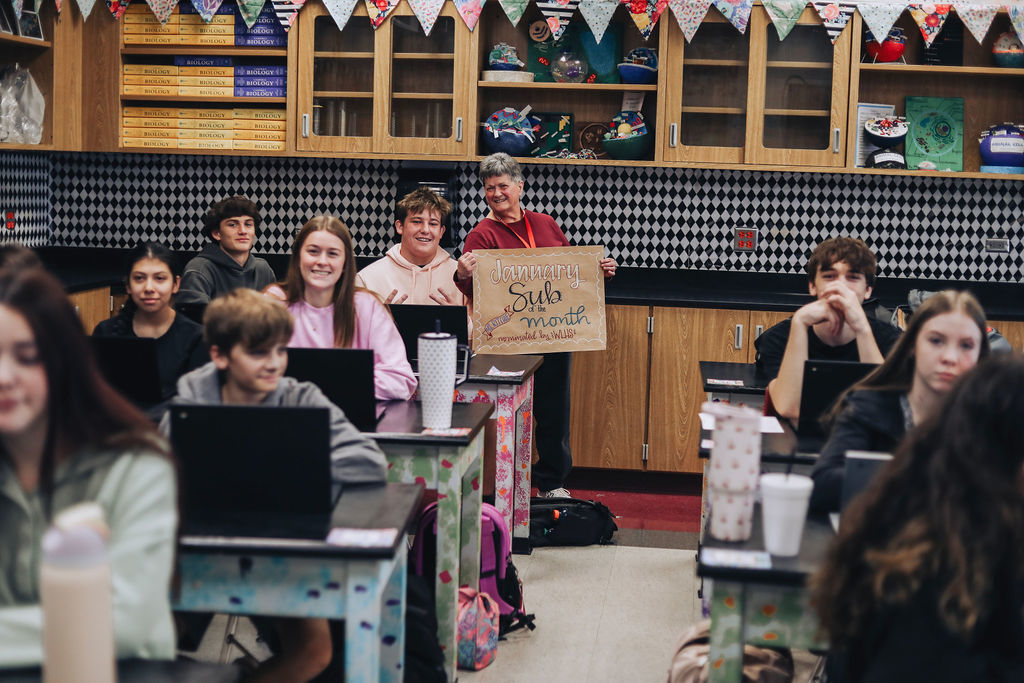 Students smile from their desks as a substitute teacher stands in the center of the classroom holding a hand-lettered sign that reads “January Sub of the Month.”