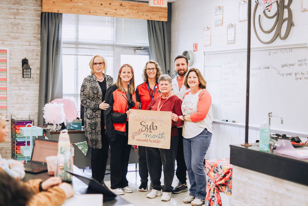 A group of school administrators and staff pose together in a classroom holding a “January Sub of the Month” sign to recognize a substitute teacher, with students seated nearby.