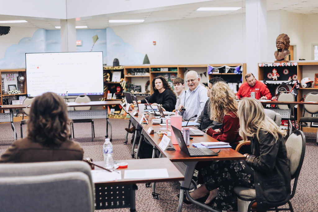 A group of adults sit around U-shaped tables in a school media center during a meeting, using laptops and papers while looking toward a presentation screen at the front of the room.