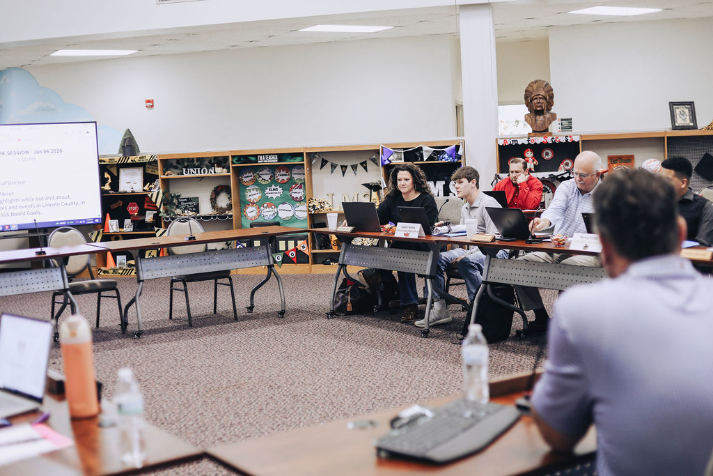 Meeting participants sit at tables in a school library space with shelves displaying school memorabilia behind them, listening and taking notes as a presentation is displayed on a large screen.