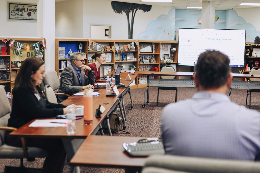 Several adults participate in a meeting in a school library setting, seated at tables with laptops and notebooks while a presentation slide is projected on a screen in front of the group.