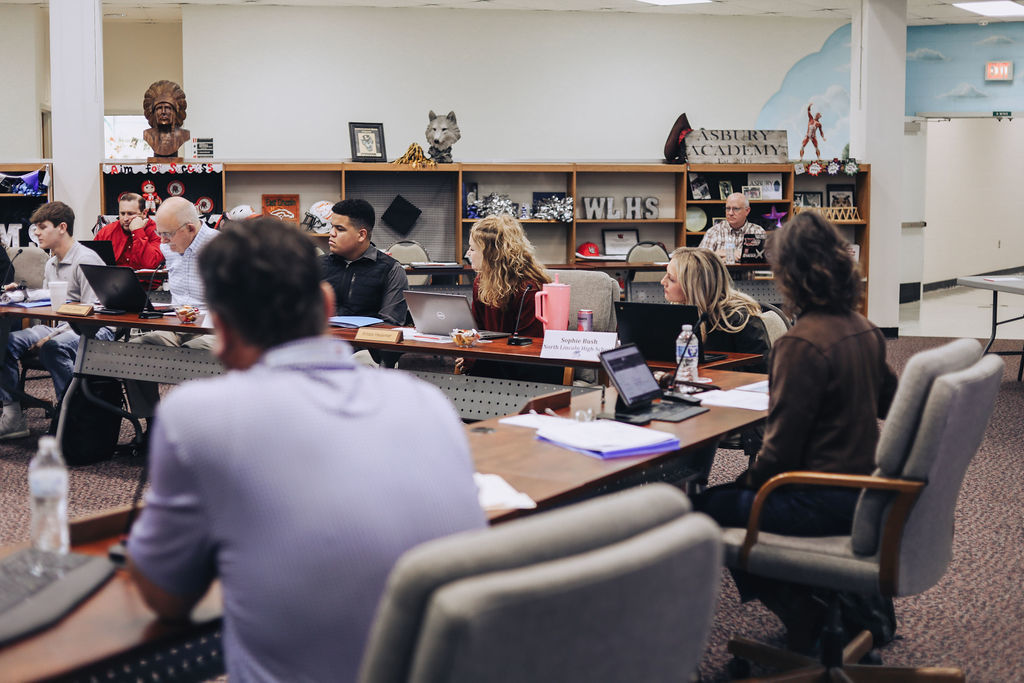 Adults seated around conference tables in a media center look toward a speaker off camera, with laptops open and school display shelves visible in the background.