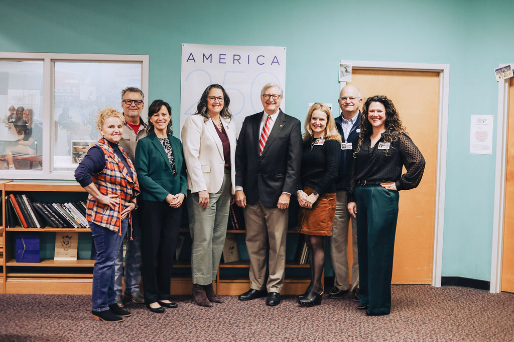 A group of district leaders and state representatives stand together for a group photo in a school media center, smiling in front of bookshelves and a sign reading “America 250 NC.”