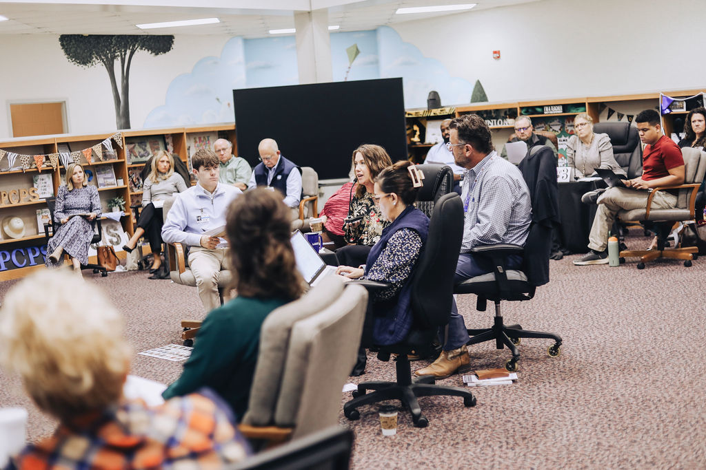 A large group of district and school leaders sit in a circle of chairs in a school media center during a meeting, engaging in discussion with laptops and printed materials visible.