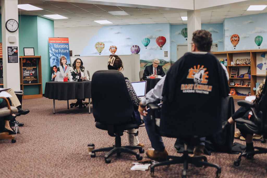 District leaders and state representatives sit at tables facing an audience during a legislative breakfast meeting in a school library, with attendees seated in rows listening and taking notes.