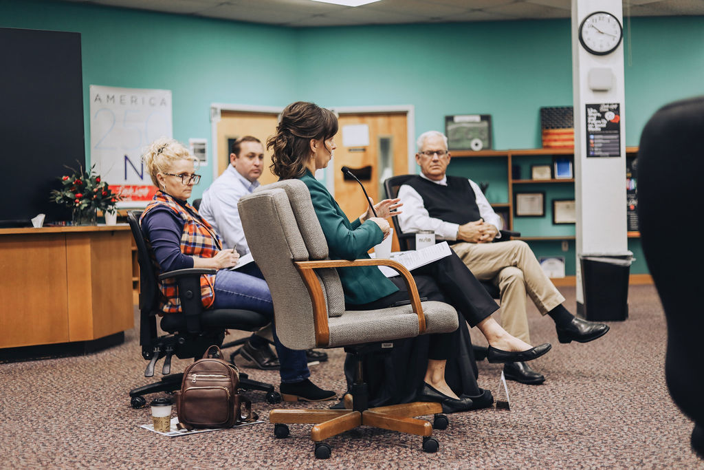 Several adults sit in rolling chairs during a meeting in a school media center, listening as one participant speaks into a microphone while others take notes.