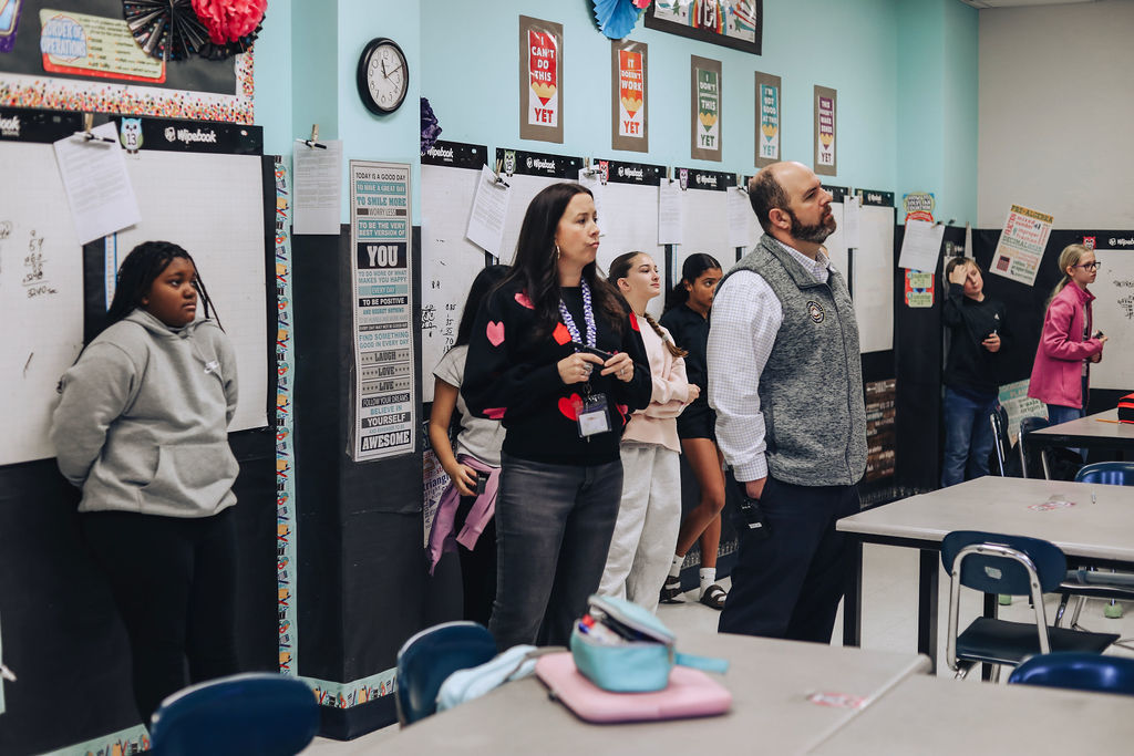 A group of students stand along the wall inside a classroom while two administrators observe and listen, with student work, posters, and whiteboards visible around the room.