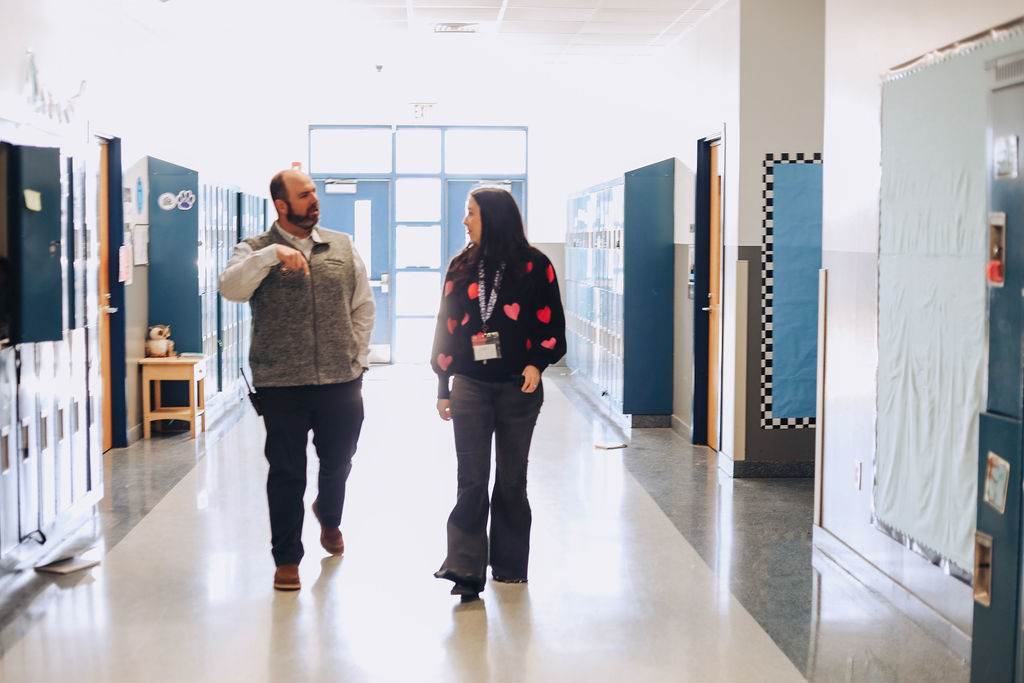 Two school administrators walk and talk down a bright school hallway lined with lockers and classroom doors, sunlight coming through glass doors at the end of the corridor.
