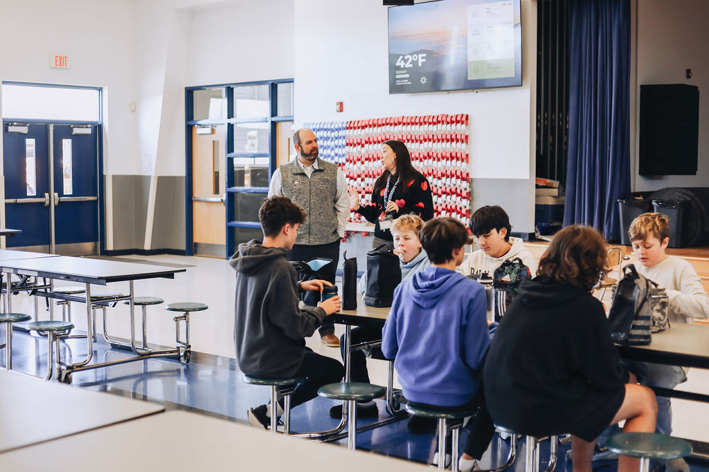 Students sit at cafeteria tables eating and talking while two administrators stand nearby observing, with a large display screen and school decor visible in the background.