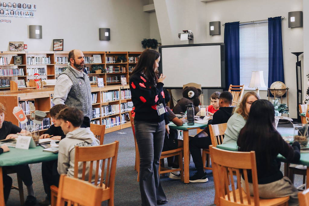 Students sit at tables in a school media center working on activities while two administrators walk through the space, bookshelves and a projection screen visible in the background.