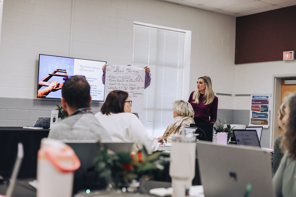 Educators sit at tables with laptops open during a professional development meeting while a presenter stands and speaks near a screen displaying a slide about the Four Disciplines of Execution.