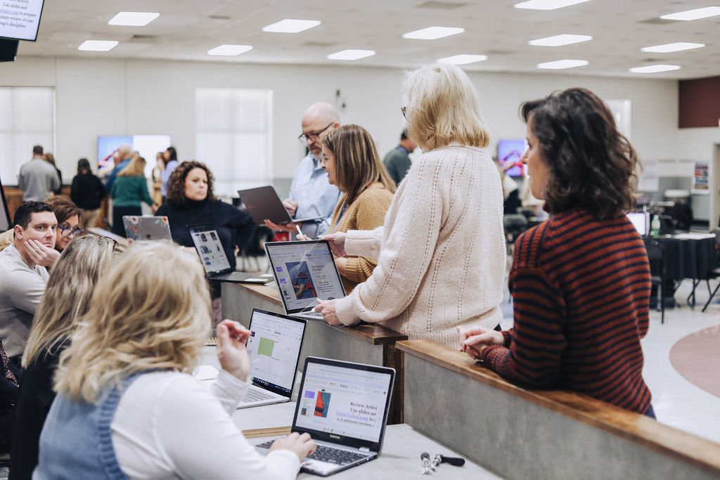 Educators gather around high tables in a large multipurpose room working on laptops and reviewing materials together during a collaborative professional learning session, with multiple groups visible in the background.
