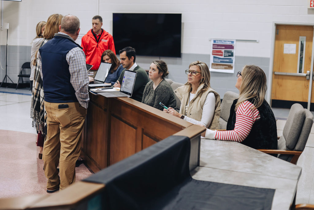Several staff members sit at a long table using laptops while a small group of colleagues stand nearby discussing information during a collaborative meeting in a large room.
