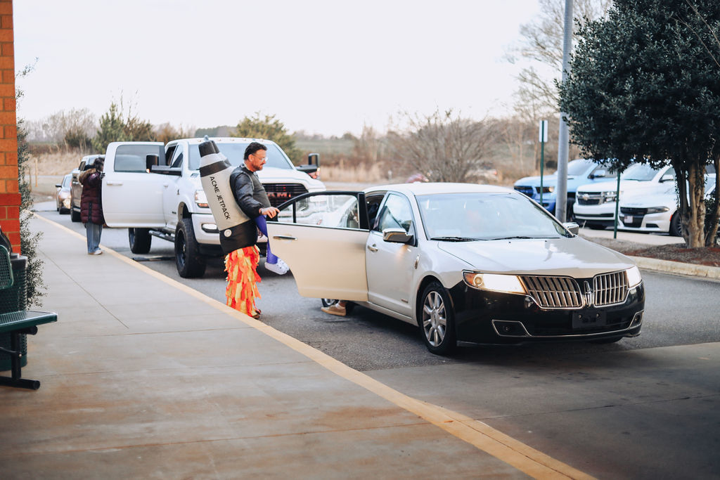 An adult in a rocket costume holds hands with a young student on the sidewalk near the school entrance while cars line up for drop-off. Another adult and child stand farther back near the building.