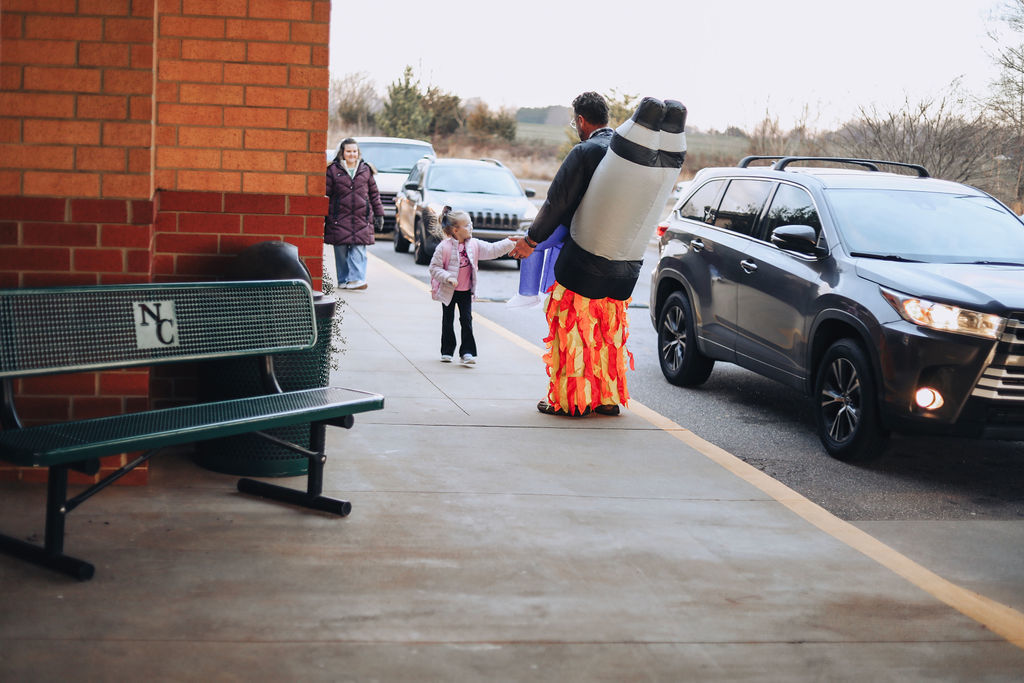 An adult in a rocket costume opens the back door of a silver sedan in the school car rider line while another vehicle waits behind. A student is partially visible entering the car, and the school building is along the sidewalk.