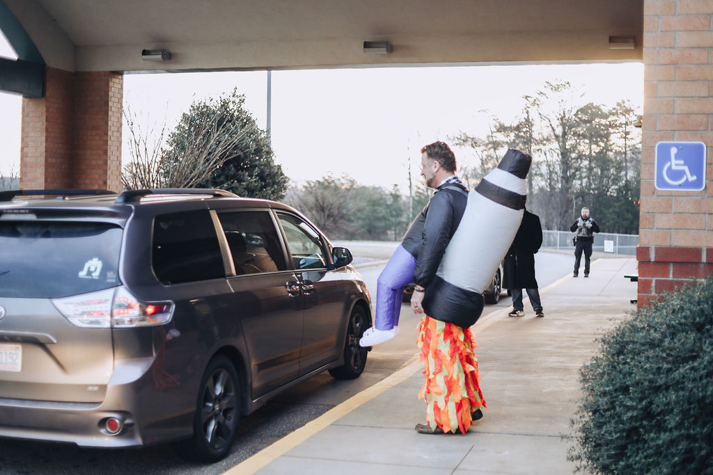 An adult wearing a playful rocket costume with flame-colored streamers stands under a school entrance canopy, holding hands with a student wearing a backpack as they walk along the sidewalk during morning arrival. Other students and adults are visible in the background near the curb.