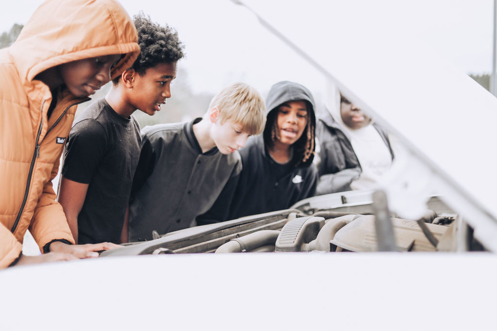 Several students lean over the open hood of a car, closely examining the engine while an adult stands nearby providing guidance.