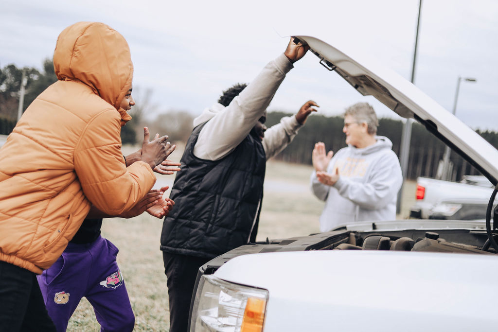 Students clap and celebrate as one student lifts the car hood closed while an adult applauds in the background in an outdoor parking area.