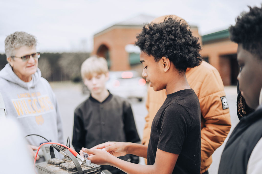 A group of middle school students stand in a parking lot while one student connects jumper cables to a car battery as an adult watches and supervises.