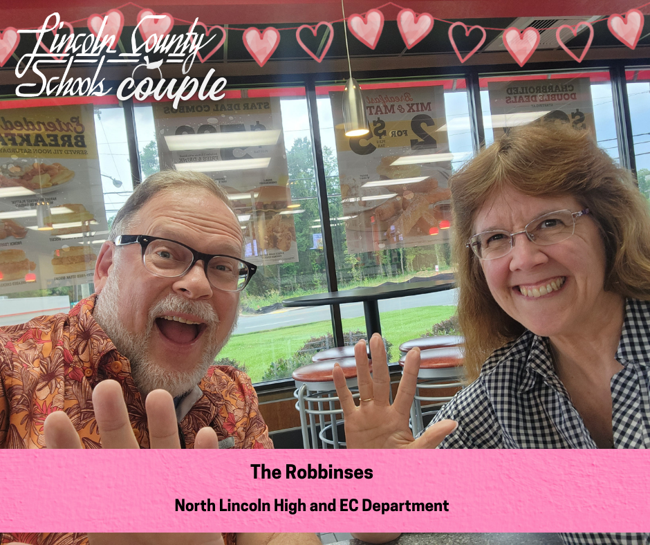 A smiling couple sits at a table inside a fast-food restaurant, both raising one hand in a friendly wave toward the camera. The person on the left wears glasses and a patterned orange shirt, and the person on the right wears glasses and a black-and-white checkered top. Large windows behind them show menu posters and a view of greenery outside. Heart graphics and the text “Lincoln County Schools couple” appear across the top. A pink banner at the bottom reads “The Robbinses” and “North Lincoln High and EC Department.”