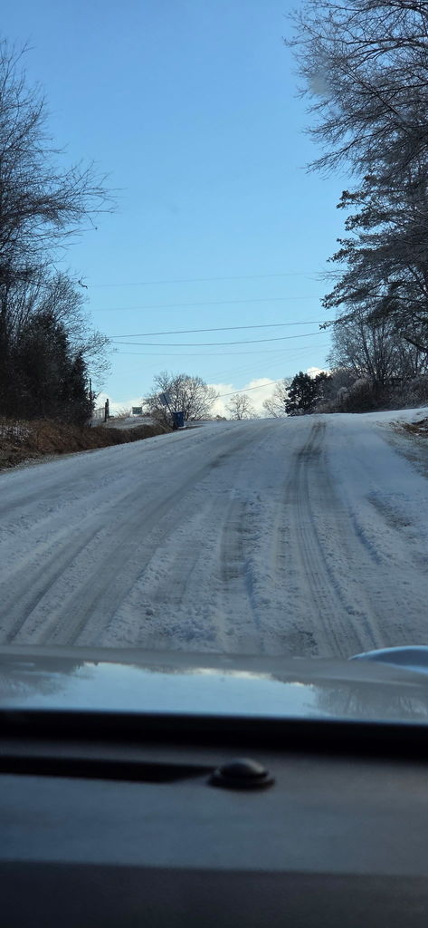 Driver’s view of a rural road covered in snow and ice, with tire tracks visible, trees lining both sides, and clear blue sky overhead.