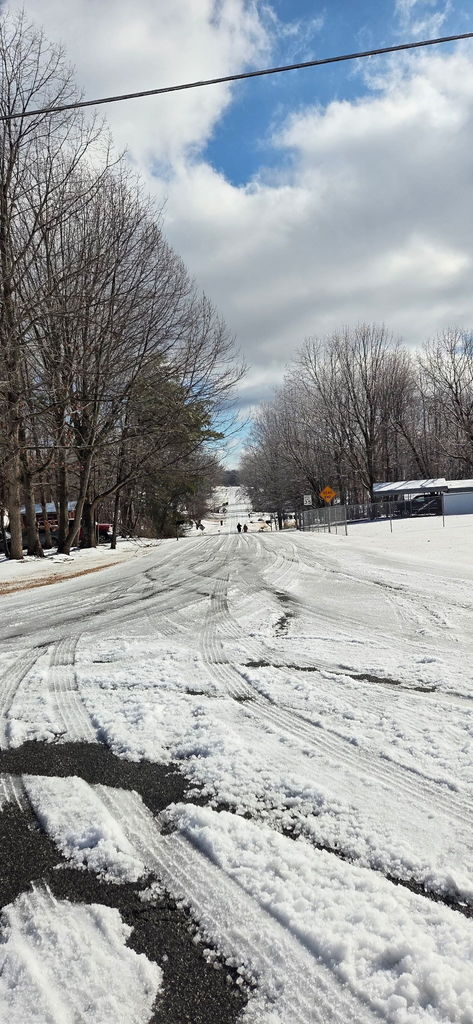 Snow-covered residential street with visible tire tracks, bare trees, road signs, and several people walking in the distance under a partly cloudy sky.