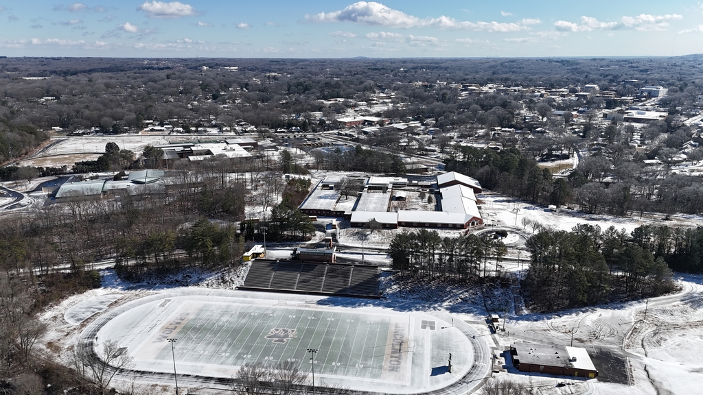 Aerial view of a snow-covered high school campus with a football stadium, track, school buildings, and surrounding neighborhood under a partly cloudy winter sky.