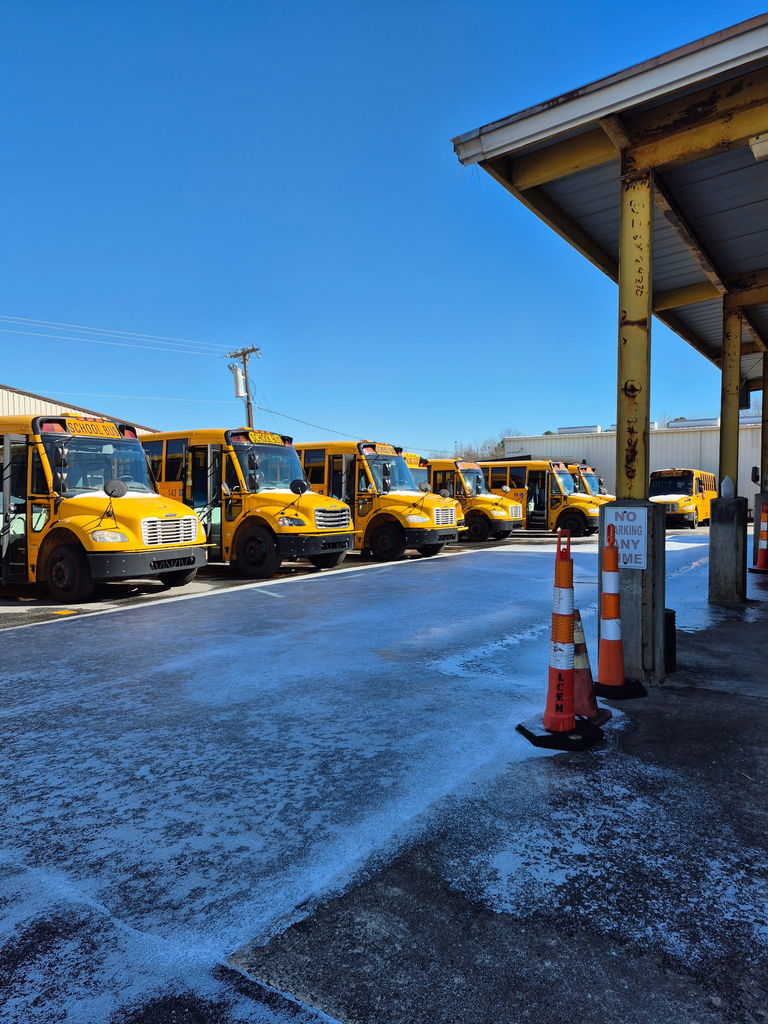 Multiple yellow school buses parked in a row at the transportation facility with ice visible on the pavement and safety cones near a covered loading area.