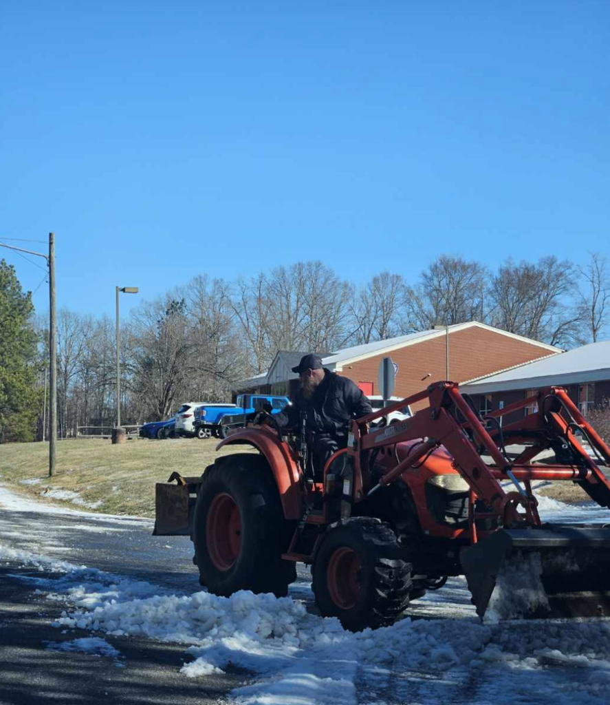 Facilities employee operating a tractor with a front blade to push slush and ice off a school driveway near parked vehicles and a brick school building.