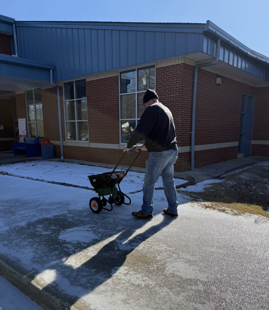 Facilities employee pushing a salt spreader across an icy sidewalk beside a brick school building to treat slippery walking areas.