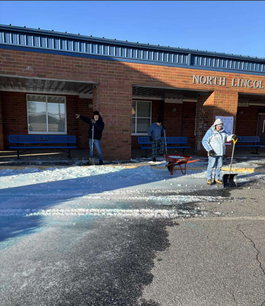 Several facilities employees shovel and spread ice melt in front of a brick school entrance while a wheelbarrow and benches sit nearby.