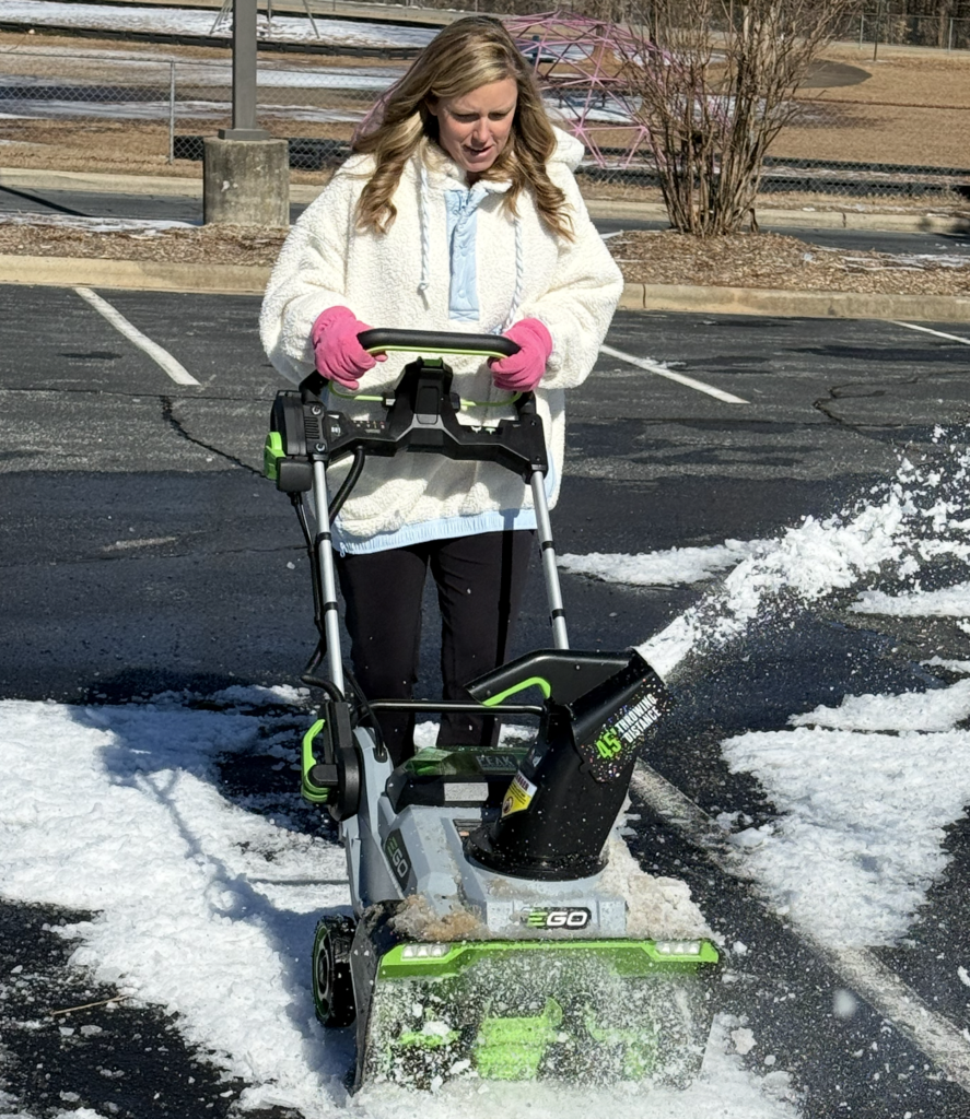 School staff member wearing pink gloves operates a small snow blower to clear snow and slush from a school parking lot.
