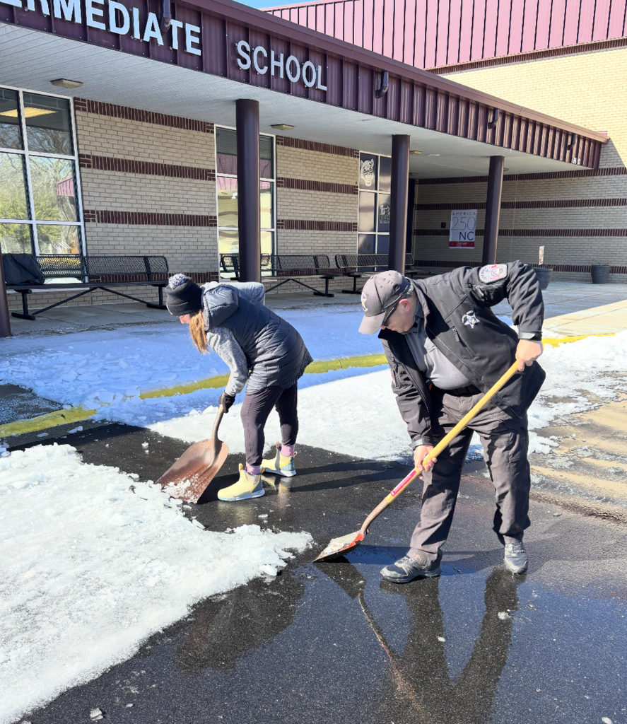 Two school staff members use shovels to clear ice and slush from the driveway in front of an intermediate school entrance with benches and columns visible.