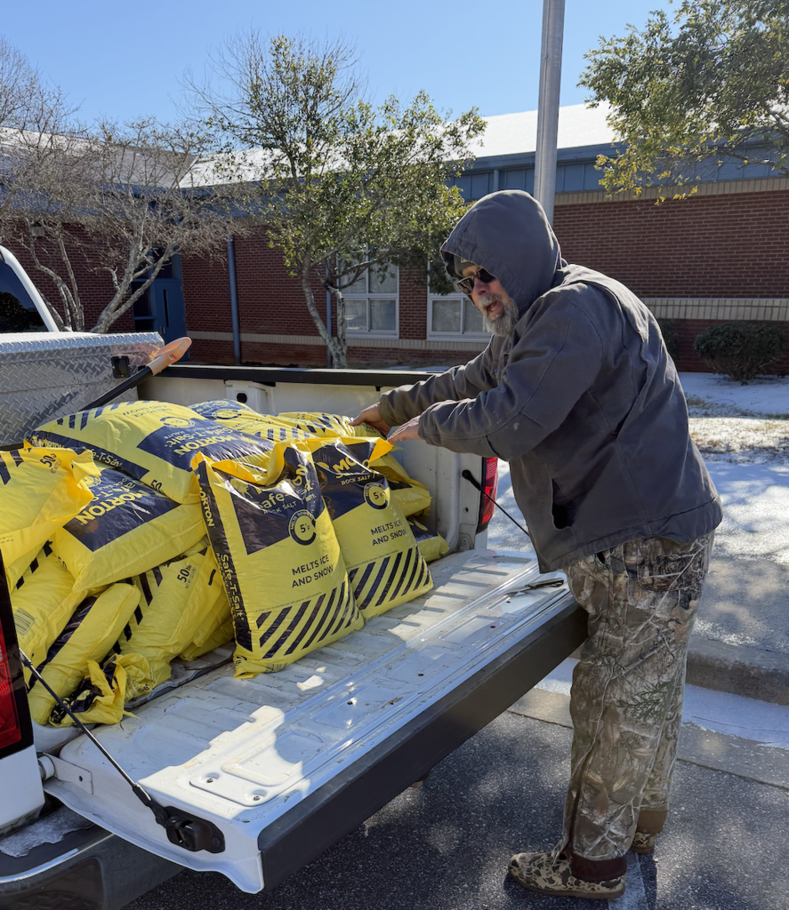 Facilities employee standing at the back of a pickup truck unloading large yellow bags of ice melt and snow treatment in a school parking area.